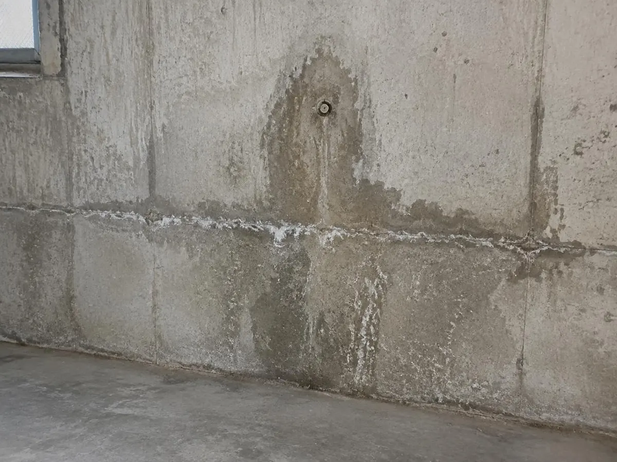Betonkellerwand mit dunklen Feuchtestellen an einer Arbeitsfuge und um eine Schalungsankerstelle, mit weißen Ablagerungen