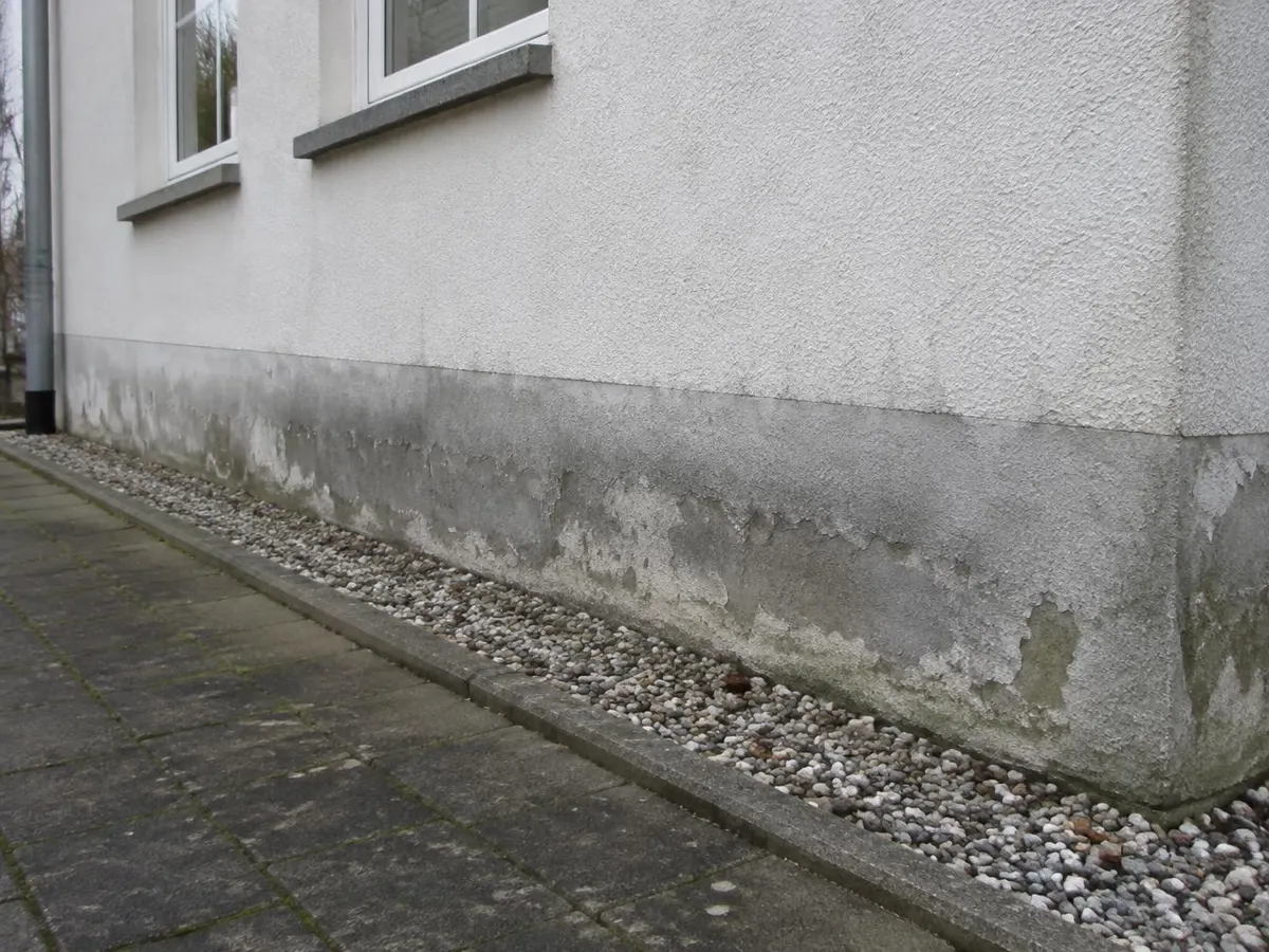 Hausfassade mit dunklem, fleckigem Sockelputz und abplatzender Beschichtung oberhalb eines Kiesstreifens