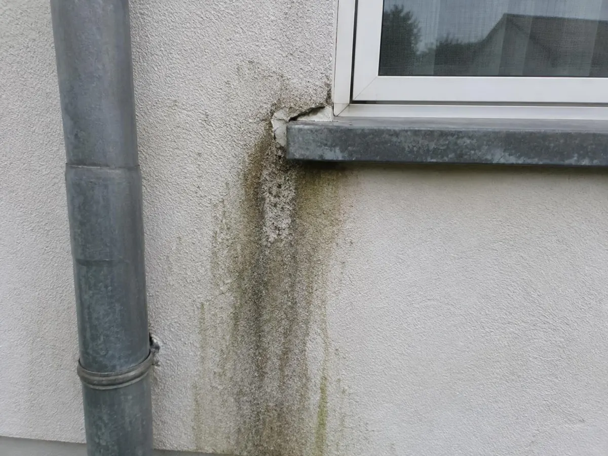 Verputzte Fassade mit senkrechten dunklen Laufspuren unterhalb einer Fensterbank neben einem Fallrohr